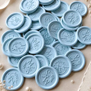 Puede incluir: Un conjunto de sellos de cera azul claro con iniciales en relieve y detalles florales. Los sellos redondos están dispersos, lo que sugiere su uso en invitaciones o papelería. También se ven perlas y flores delicadas.