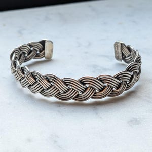 May include: A silver-toned cuff bracelet featuring a continuous braided or woven metal pattern. The design consists of multiple thin, parallel strands intertwined to create a textured, rope-like appearance with smooth, solid end caps.