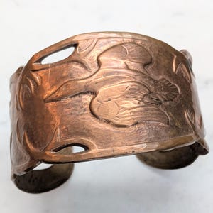 May include: A wide copper cuff bracelet featuring a raised relief design of a bird in flight. The metal has a warm, reddish-brown metallic finish with cut-out sections along the top and bottom edges, creating a detailed, textured surface.
