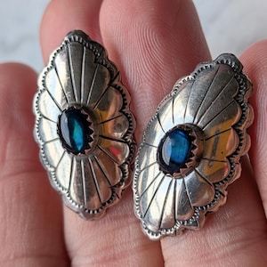 May include: A pair of oval-shaped silver earrings featuring a sunburst pattern with scalloped edges. Each earring is centered with a small, smooth, dark blue oval stone set in a serrated bezel. The silver has a polished finish with dark oxidation.