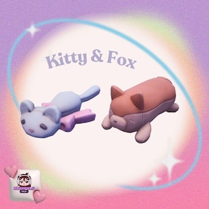 May include: Two plush animal toys: a light blue cat and a pink and brown fox. The cat is lying down with a pink bow. The fox is also lying down. The text "Kitty & Fox" is above the toys.