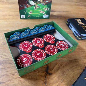 Pode incluir: Uma caixa de cart&atilde;o verde contendo fichas de casino azuis e vermelhas. As fichas vermelhas t&ecirc;m n&uacute;meros e s&iacute;mbolos brancos. Uma caixa de jogo com um rato de desenho animado a jogar cartas est&aacute; no fundo. Uma caixa preta com a palavra "Ratjac" tamb&eacute;m &eacute; vis&iacute;vel.