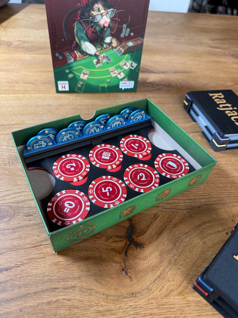 Pode incluir: Uma caixa de cart&atilde;o verde contendo fichas de casino azuis e vermelhas. As fichas vermelhas t&ecirc;m n&uacute;meros e s&iacute;mbolos brancos. Uma caixa de jogo com um rato de desenho animado a jogar cartas est&aacute; no fundo. Uma caixa preta com a palavra "Ratjac" tamb&eacute;m &eacute; vis&iacute;vel.