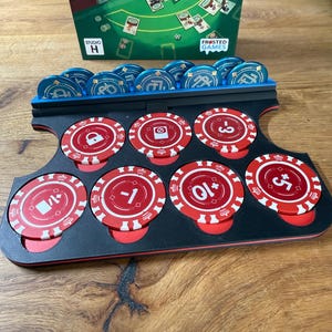 Pode incluir: Um suporte preto e vermelho para fichas de poker com fichas azuis e vermelhas. As fichas t&ecirc;m v&aacute;rios s&iacute;mbolos e n&uacute;meros impressos. A caixa do jogo no fundo tem o texto "Frosted Games" e "Studio H".