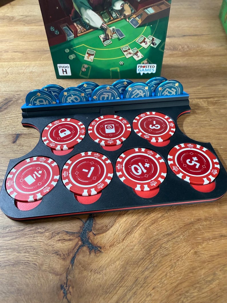 Pode incluir: Um suporte preto e vermelho para fichas de poker com fichas azuis e vermelhas. As fichas t&ecirc;m v&aacute;rios s&iacute;mbolos e n&uacute;meros impressos. A caixa do jogo no fundo tem o texto "Frosted Games" e "Studio H".