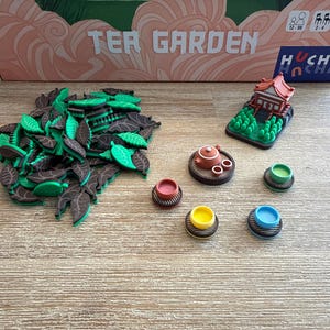 Könnte beinhalten: Ein Brettspiel namens "Tea Garden" mit Spielsteinen. Die Spielsteine umfassen grüne und braune Blattformen, Miniatur-Teesets und farbige Schalen. Ein rotes und braunes Miniaturgebäude ist ebenfalls enthalten.