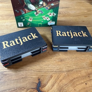 Könnte beinhalten: Zwei schwarze Kartenhalter mit dem Wort "Ratjack" in goldfarbenen Buchstaben werden gezeigt. Die Spielbox zeigt eine Cartoon-Ratte beim Kartenspielen. Das Spiel heißt "Ratjack" und stammt von Frosted Games.
