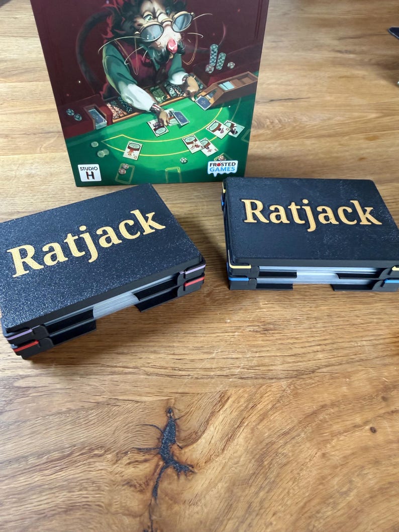 Pode incluir: Dois porta-cart&otilde;es pretos com a palavra "Ratjack" em letras douradas s&atilde;o exibidos. A caixa do jogo apresenta um rato de desenho animado jogando cartas. O jogo chama-se "Ratjack" e &eacute; da Frosted Games.