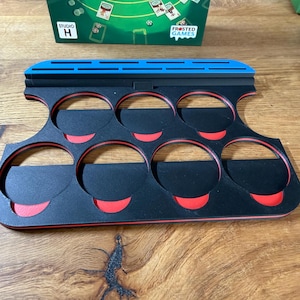 Pode incluir: Um suporte de pe&ccedil;as de jogo preto e vermelho com seis ranhuras circulares. O suporte tem uma borda superior azul e foi projetado para segurar pe&ccedil;as de jogo. As palavras "FROSTED GAMES" s&atilde;o vis&iacute;veis em uma caixa de jogo ao fundo.