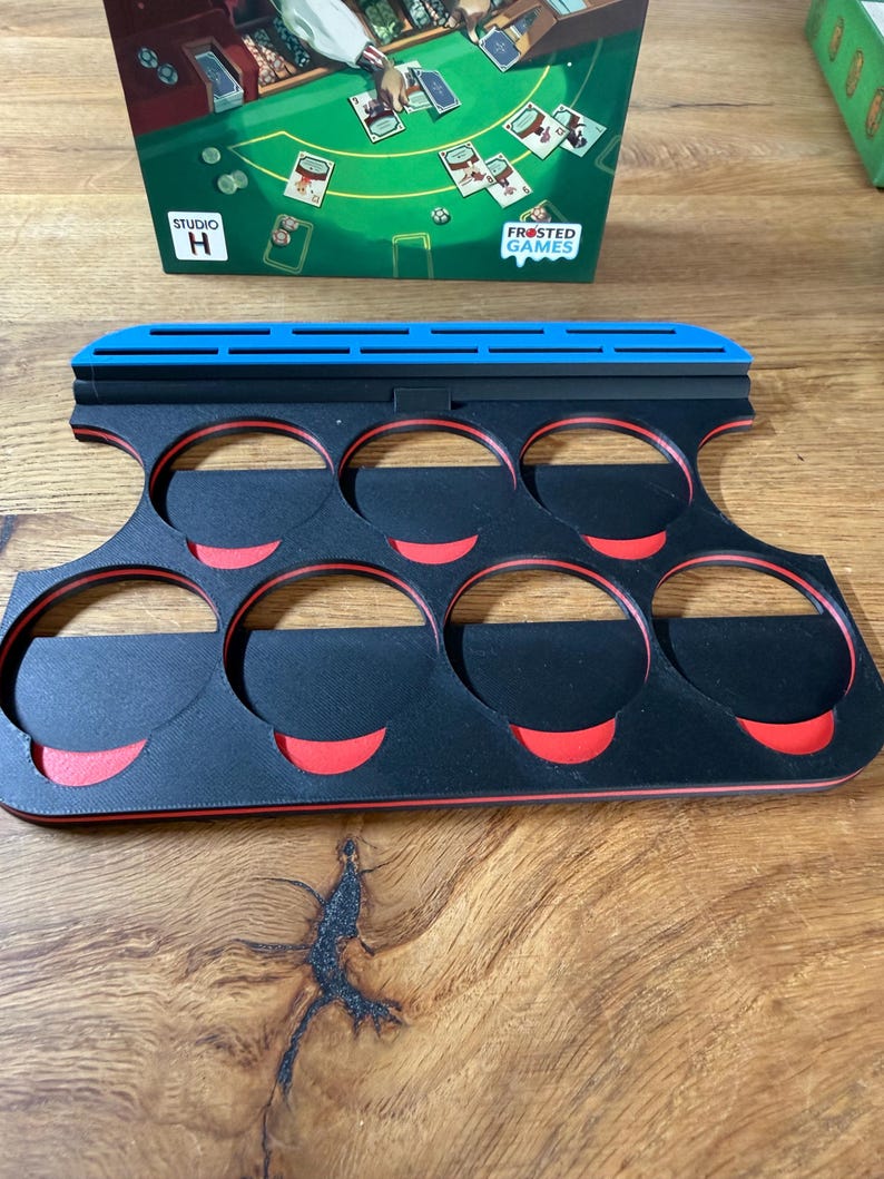 Pode incluir: Um suporte de pe&ccedil;as de jogo preto e vermelho com seis ranhuras circulares. O suporte tem uma borda superior azul e foi projetado para segurar pe&ccedil;as de jogo. As palavras "FROSTED GAMES" s&atilde;o vis&iacute;veis em uma caixa de jogo ao fundo.