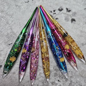 Op de afbeelding: Een set van zeven taps toelopende pennen in een waaieropstelling, elk met een behuizing van heldere hars gevuld met kleurrijke glitters, metallic vlokken en kleine goudkleurige roosversieringen in groen, paars, roze, goud, blauw en magenta tinten.