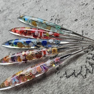 Può includere: Cinque penne in resina trasparente disposte a ventaglio, ognuna contenente elementi unici incorporati tra cui fiori secchi, scaglie di foglia d'oro e piccole forme di farfalla in vari colori come blu, rosso, bianco e rosa con accenti metallici.