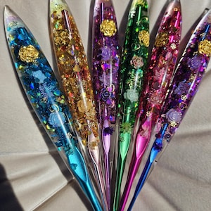Può includere: Sei forcine per capelli colorate con corpi in resina trasparente riempiti di glitter e dettagli floreali. Le forcine presentano una varietà di colori, tra cui blu, oro, viola, verde, rosa e un viola intenso. Ogni forcina ha una base metallica.
