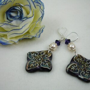 May include: A pair of handcrafted earrings featuring square, dark purple ceramic pendants with a teal and gold floral design. Each earring has a pearl bead, purple crystal accents, and silver-tone ear wires. A fabric flower with blue trim is in the background.