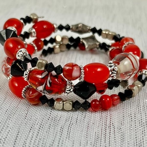 May include: A multi-strand bracelet featuring red, black, and silver beads. The bracelet includes various bead shapes, including round, faceted, and decorative metal accents. The beads are strung on a flexible wire, creating a wrap-around design.