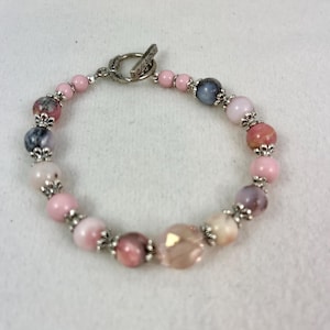 May include: A beaded bracelet featuring a variety of pink, purple, and gray beads. The bracelet includes a mix of round and faceted beads, with silver-colored metal accents and a toggle clasp. The beads vary in size and shade, creating a delicate and feminine accessory.