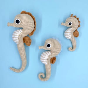 Op de afbeelding: Drie gehaakte zeepaardjesfiguren in verschillende maten. De zeepaardjes zijn voornamelijk beige met witte buiken, bruine vinnen en zwarte ogen. Eén zeepaardje heeft een blauwe vin. De achtergrond is effen lichtblauw.
