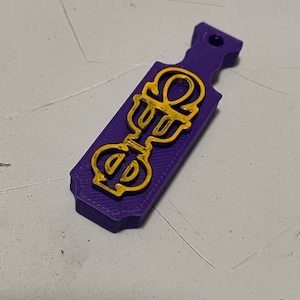 May include: A purple paddle-shaped pendant with a gold-colored ankh symbol. The pendant has a hole at the top for hanging. The ankh symbol is a stylized cross with a loop at the top, often associated with ancient Egyptian culture.