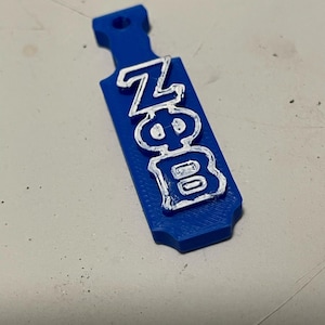 May include: A blue paddle-shaped item with the Greek letters ZFB in white. The paddle has a handle with a hole at the top. The letters are raised and outlined in white, creating a 3D effect. The item is likely a decorative or symbolic piece.