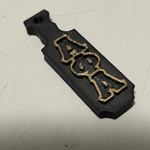 May include: Black paddle-shaped pendant with a gold-colored design featuring the letters "AOA". The pendant has a hole at the top for hanging. The design is raised and has a textured appearance.