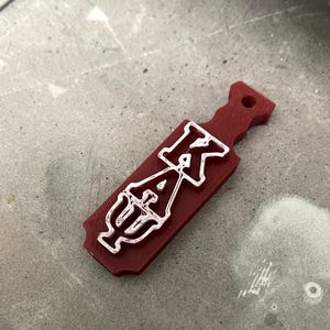 May include: A maroon paddle-shaped item with a white outline of the Greek letters Kappa, Alpha, and Psi. The item has a hole at the top and is set against a gray surface.