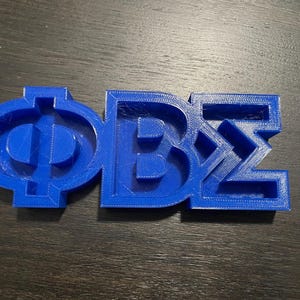 Peut inclure: Objet bleu imprimé en 3D représentant les lettres grecques ΦΒΣ. Les lettres sont reliées et présentent un aspect texturé, probablement dû au processus d'impression. L'objet est posé sur une surface en bois sombre.