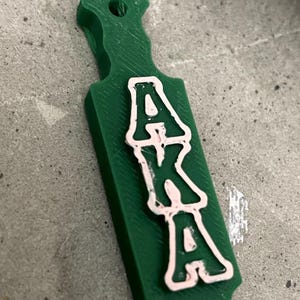 Puede incluir: Un llavero verde en forma de paleta con las letras "AKA" en blanco con un contorno rosa. El llavero tiene un asa con un agujero para sujetarlo a un llavero. Las letras están apiladas verticalmente en la paleta.
