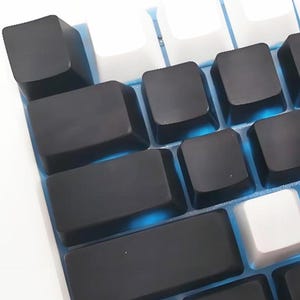 Może przedstawiać: Zbliżenie klawiatury mechanicznej z czarnymi i białymi klawiszami. Klawisze są ułożone w siatkę, z mieszanką czarnych i białych klawiszy. Klawiatura ma niebieskie podświetlenie.