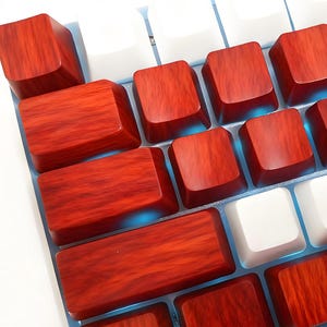 Puede incluir: Primer plano de un teclado mecánico con teclas personalizadas. Las teclas alternan entre un patrón de vetas de madera en tonos rojos y blancos. El teclado tiene un marco blanco y retroiluminación azul.