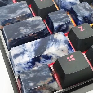 May include: Close-up of a mechanical keyboard with custom keycaps. The keycaps are made of a dark blue and white stone, with black keys in between. The keyboard has red LED lighting.