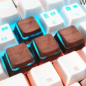 May include: Close-up of a mechanical keyboard with white keys and brown wooden keycaps. The WASD keys are replaced with wooden keycaps. The keys are backlit with blue and orange lights. The text "WASD" is visible.
