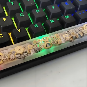 May include: A custom mechanical keyboard with black keycaps and a unique spacebar. The spacebar is clear and filled with an assortment of gold and silver gears. The keyboard has colourful backlighting.