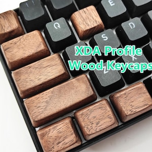 Puede incluir: Primer plano de un teclado con teclas de madera. Las teclas son de color marrón cálido, con vetas de madera visibles. El teclado también tiene teclas negras. El texto en la imagen dice "XDA Profile Wood Keycaps."