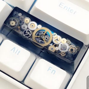 Puede incluir: Una tecla de teclado personalizada con un diseño detallado de engranajes mecánicos en tonos dorados, plateados y bronce, encapsulados en resina transparente. La tecla se encuentra entre teclas de teclado blancas, con las teclas Enter, Alt y Fn visibles.