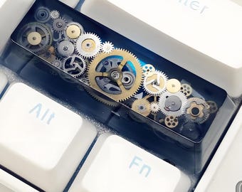Könnte beinhalten: Eine individuelle Tastaturkappe mit einem detaillierten Design mechanischer Zahnräder in Gold-, Silber- und Bronzetönen, eingebettet in transparentem Harz. Die Kappe befindet sich zwischen weißen Tastaturtasten, wobei die Tasten Enter, Alt und Fn sichtbar sind.