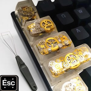 以下が含まれることがあります： カスタムキーキャップ付きのメカニカルキーボードのクローズアップ。キーキャップは透明な樹脂製で、金と銀の時計の歯車が収められています。「Esc」のロゴとキーキャッププーラーも表示されています。