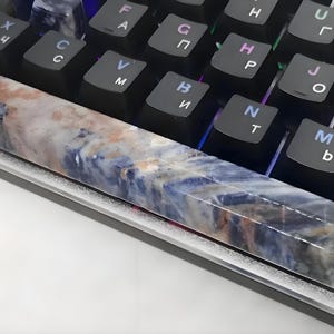 May include: A custom artisan keyboard spacebar keycap with a unique design. The keycap features a marbled pattern with shades of blue, brown, and white. It rests on a black mechanical keyboard with white lettering.