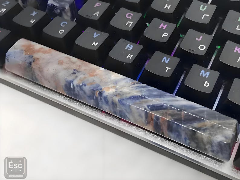 May include: A custom artisan keyboard spacebar keycap with a unique design. The keycap features a marbled pattern with shades of blue, brown, and white. It rests on a black mechanical keyboard with white lettering.