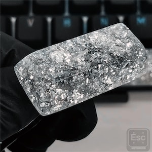 May include: A rectangular, clear resin keycap filled with silver flakes. The keycap is held by a gloved hand, with a black keyboard in the background. The keycap has a shiny, reflective surface.