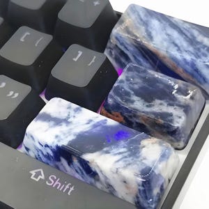 May include: Close-up of a keyboard with custom keycaps. The keycaps are made of a blue and white stone-like material. The keyboard keys are black and gray. The Shift key is visible, along with other keys.