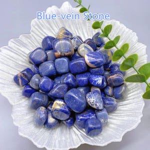 May include: A collection of polished blue-vein stones, with a mix of blue and white veining, displayed in a decorative, white, petal-shaped dish. The stones are smooth and rounded, with a glossy finish. The text "Blue-vein Stone" is visible.