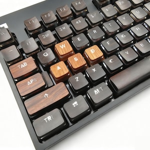 Pu&ograve; includere: Primo piano di una tastiera di computer nera con tasti in legno. I tasti per W, A, S e D sono in legno chiaro. Altri tasti sono marrone scuro. La tastiera ha la lettera "G" nell'angolo in alto a sinistra.