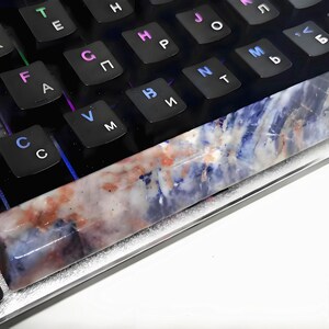 May include: A rectangular, multi-colored gemstone keycap rests on a black mechanical keyboard. The keycap features a blend of blue, white, and reddish-brown hues. The keyboard has black keys with white and multi-colored lettering.