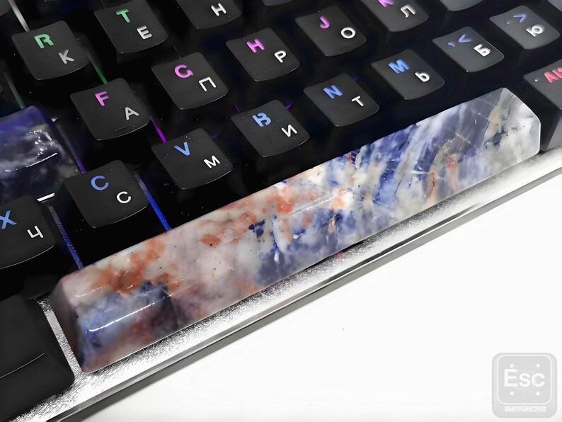 May include: A rectangular, multi-colored gemstone keycap rests on a black mechanical keyboard. The keycap features a blend of blue, white, and reddish-brown hues. The keyboard has black keys with white and multi-colored lettering.