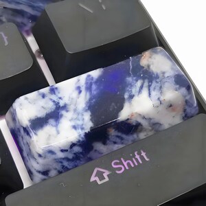 May include: Close-up of a custom keyboard with a unique artisan keycap. The keycap is a rectangular shape with a marbled design of white, blue, and hints of pink. The keyboard keys are black, with the word "Shift" visible on one key.