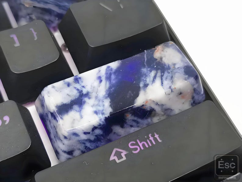 May include: Close-up of a custom keyboard with a unique artisan keycap. The keycap is a rectangular shape with a marbled design of white, blue, and hints of pink. The keyboard keys are black, with the word "Shift" visible on one key.