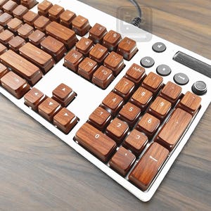 Pu&ograve; includere: Una tastiera per computer con tasti in legno e una cornice argentata. I tasti sono di un caldo colore marrone e la tastiera ha pulsanti di controllo multimediale e una manopola del volume. La tastiera &egrave; su una superficie di legno.