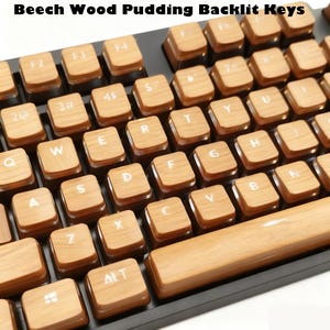 May include: A mechanical keyboard featuring custom beech wood pudding keycaps with a light brown wood grain texture. The keys have white translucent lettering for backlighting, including standard QWERTY layout characters and function keys.