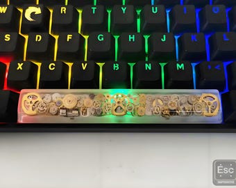 Könnte beinhalten: Eine mechanische Tastatur mit schwarzen Tasten und einer individuellen Leertaste. Die Leertaste ist transparent mit eingebetteten goldfarbenen Zahnrädern und anderen Uhrwerksteilen. Die Tastatur hat eine Regenbogen-Hintergrundbeleuchtung.