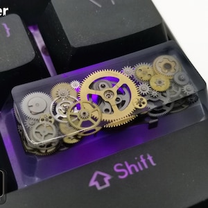 May include: A custom Enter keycap for a mechanical keyboard, featuring a clear resin design filled with various sizes of gold and silver gears. The keycap is set on a black keyboard with purple backlighting.